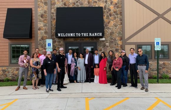 Group of people standing outside a building with a "Welcome to the Ranch" sign, some wearing uniforms, in front of a stone and wood facade, with reserved parking signs nearby.