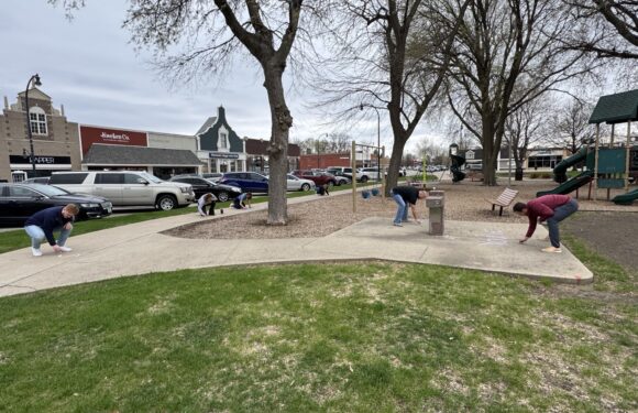 Six people are cleaning a playground area, sweeping and picking up trash around the swings and slide. There are parked cars and shops in the background.