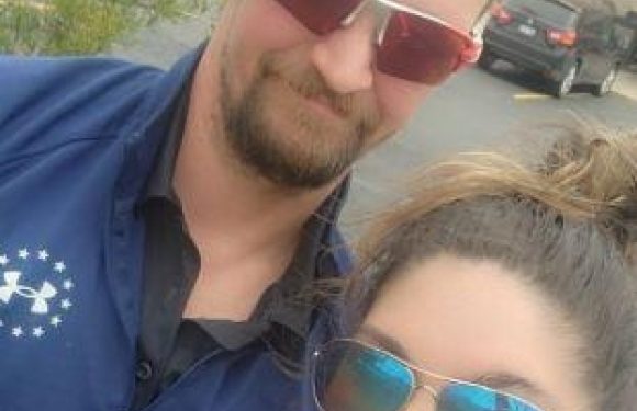 A man and woman are taking a selfie outdoors, both wearing sunglasses and smiling. The man is in a navy blue shirt and the woman has curly hair pulled back.