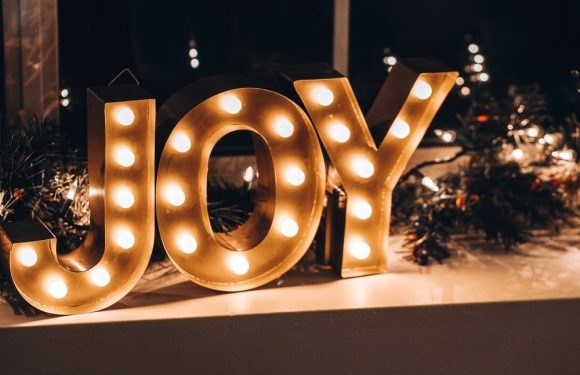 Illuminated gold "JOY" sign with round bulbs, decorated with greenery and Christmas lights, displayed indoors in a festive setting.