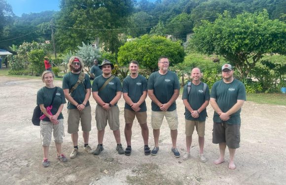 A group of seven people wearing dark green T-shirts standing outdoors on a gravel area, with green trees and hills in the background on a clear, sunny day.