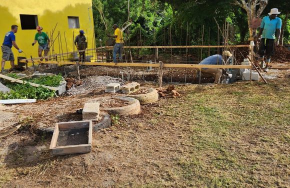 Several people are working on a construction site outside a yellow building, with trees and blue sky in the background. They are wearing casual clothing, some with hats.