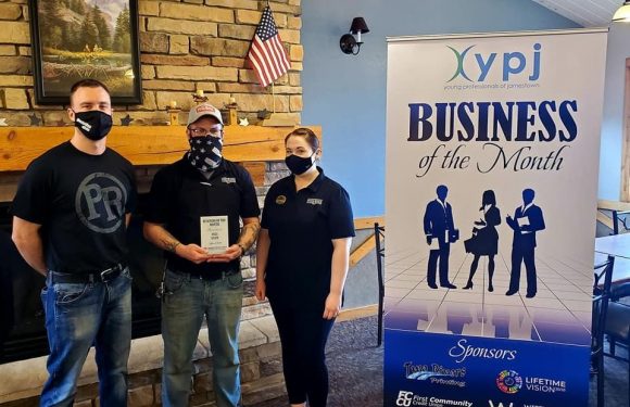 Three people, wearing black and navy clothing with masks, stand in a room near a brick fireplace, holding an award, beside a banner for YPJ Business of the Month.