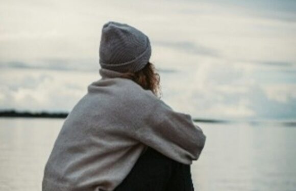 Person sitting on a dock by the water, wearing a gray sweater, a gray beanie, black pants, and gray sneakers, gazing at the cloudy sky and water.