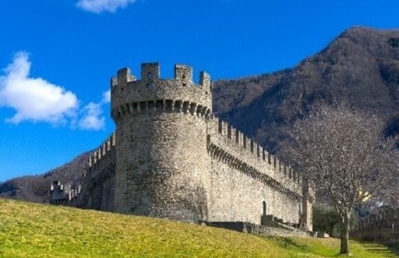 A stone castle with battlements and a round tower on a grassy hill, set against a backdrop of mountains and a bright blue sky with some clouds.