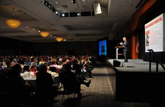 A speaker in a suit stands at a podium on a stage, addressing a large seated audience in a banquet hall with high ceilings and chandeliers.