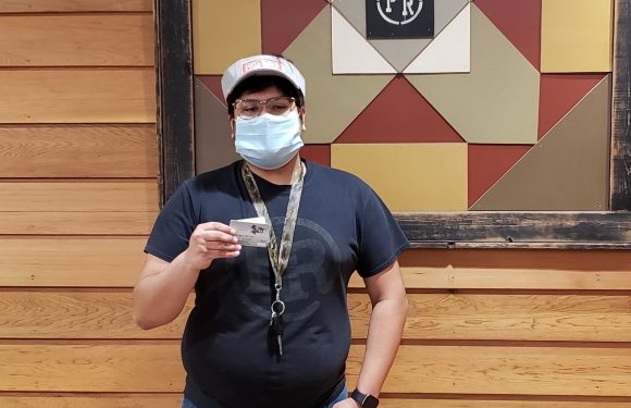 A person wearing a face mask, glasses, and a cap holding a card, standing in front of a wooden wall with a geometric sign that says "PR".