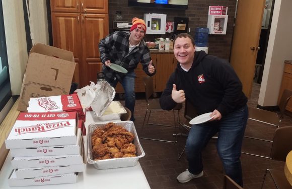 Two men are inside a room with wooden cabinets, gathered around a table with pizza boxes and fried chicken. They are smiling and giving thumbs-up gestures.