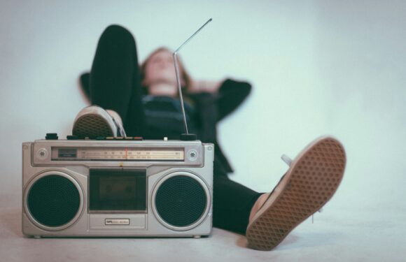 A woman relaxing with her hands behind her head, lying in front of a vintage boombox on the floor, with her feet up and slightly out of focus.