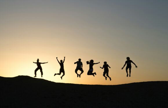 Six people are jumping in the air on a hilltop at sunset, their silhouettes visible against the sky. They appear joyful and energetic.