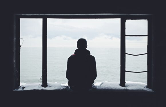 A person in a hoodie sits on a windowsill, looking out at a cloudy ocean scene through a large, framed window. The scene is dark and moody with snow on the windowsill.
