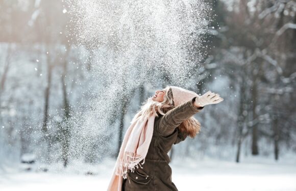 A woman dressed warmly in winter clothing, including a coat, scarf, gloves, and a hat, throws snow into the air in a snowy outdoor park.