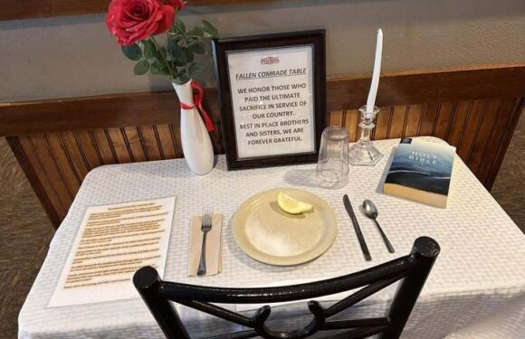 A memorial table setup with a vase of red roses, a framed tribute, a candle, a glass, a Bible, and a plate with a lemon wedge.