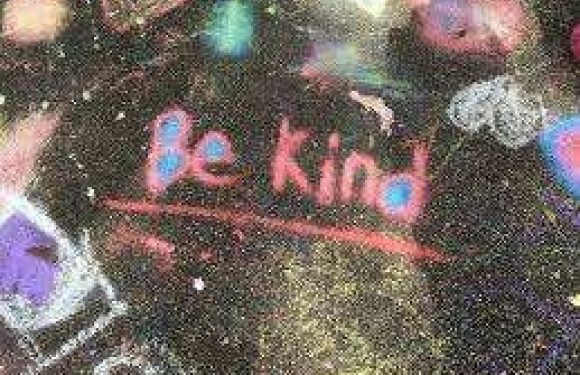 A chalkboard with "Be Kind" written in pink and red with a small heart shape, surrounded by colorful chalk smudges in a casual, creative setting.