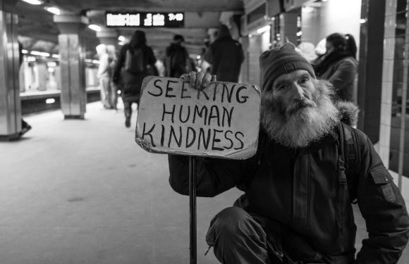 A bearded man wearing a beanie and jacket holds a cardboard sign that reads "SEEKING HUMAN KINDNESS" in a busy subway station.