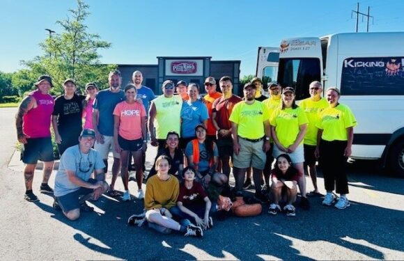 Group of people standing and sitting outside with a Pizza Ranch sign, some in bright yellow shirts, in front of a bus and a clear blue sky.