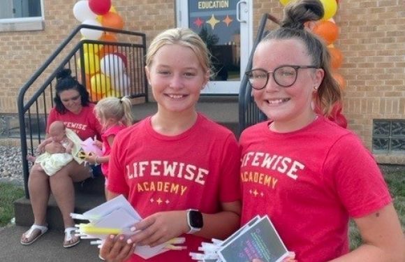 Two girls wearing red "LIFESIZE ACADEMY" t-shirts stand outside in front of a building with balloons and a sign that reads "LIFESIZE Academy Bible Based Character Education," holding pamphlets.