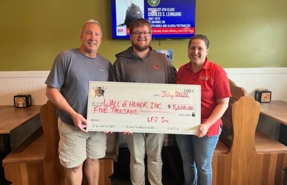 Three individuals stand together inside a restaurant, holding a large check made out to Wall of Honor Inc. for $5,000, dated July 2022. They are smiling and posing for the photo. The person on the left wears a gray T-shirt and beige shorts, the person in the middle wears a dark hoodie and glasses, and the person on the right wears a red polo shirt with a Pizza Ranch logo. In the background, a television displays a presentation with a photo and details about Charles S. Leiningg.