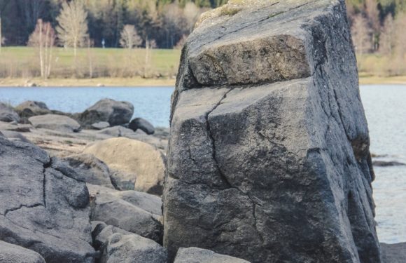 Large upright rock on a sandy beach with smaller rocks nearby, trees and a body of water in the background under a partly cloudy sky.