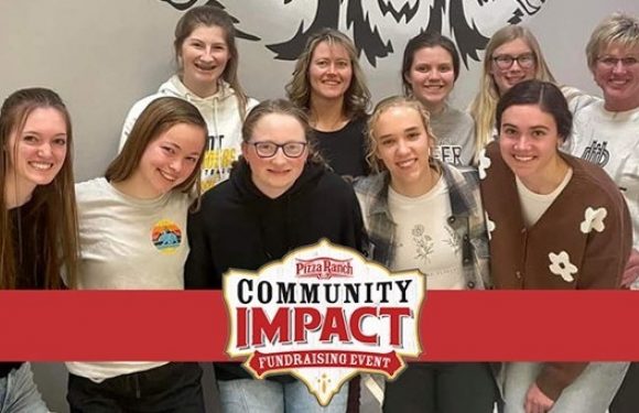 Group of nine young women smiling, some wearing hoodies and casual clothes, standing in front of a wall with a large graphic of a lion and a "Community Impact" sign.