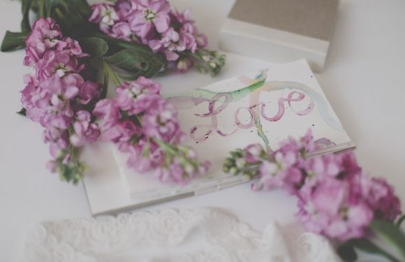 A greeting card with the word "Love" written in pastel colors, pink flowers with green leaves, and a white doily on a white surface.