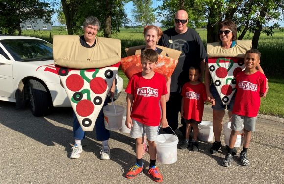 Group of people dressed as pizza slices and Pizza Ranch staff standing outdoors on a sunny day with trees and a field in the background.