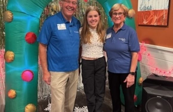 Three people, two employees in blue shirts and a girl in a white top, stand under a green balloon arch with pink and red decorations, in a decorated indoor setting.