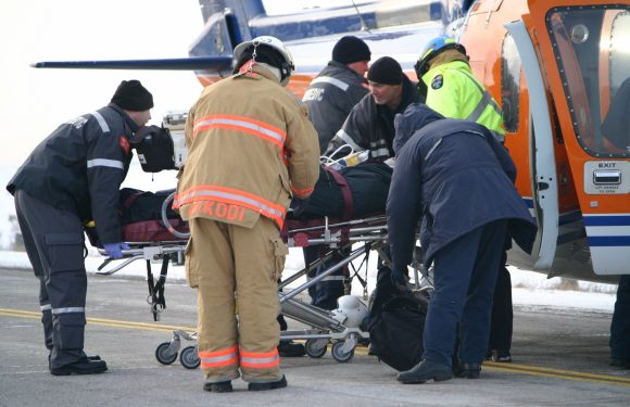 Emergency responders surround a person on a stretcher being loaded into a helicopter for transport. The crew includes firefighters, paramedics, and rescue personnel.