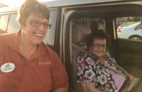 A woman in a Pizza Ranch uniform with glasses smiling next to an elderly woman sitting in a car seat, holding a book and wearing a floral shirt.