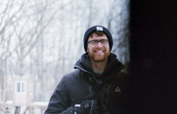 A smiling man with glasses, a beard, and a beanie, dressed in winter clothing, outdoors on a snowy day with trees in the background.