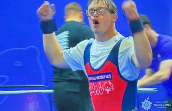 A male athlete wearing glasses and a red and blue uniform with "Special Olympics" text is celebrating with clenched fists. The background is blue with others in similar attire.