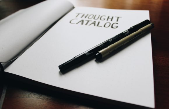 Open notebook titled "Thought Catalog" with two black and beige markers resting on it, on a wooden desk.