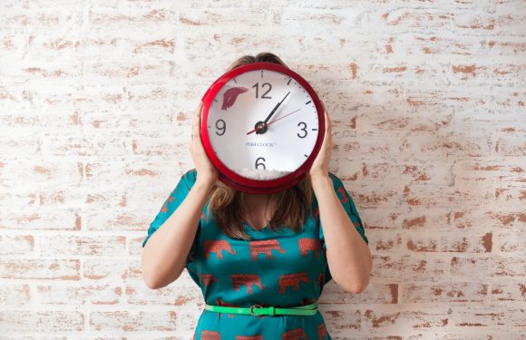 Person holding a red fish-shaped clock in front of their face, standing against a white brick wall, wearing a teal dress with orange animal prints and a light green belt.