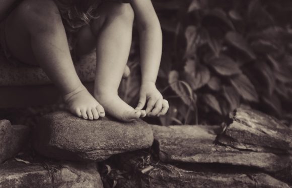 A child is sitting on a rock ledge, barefoot, with their hands reaching down. The background features leafy foliage in black and white.