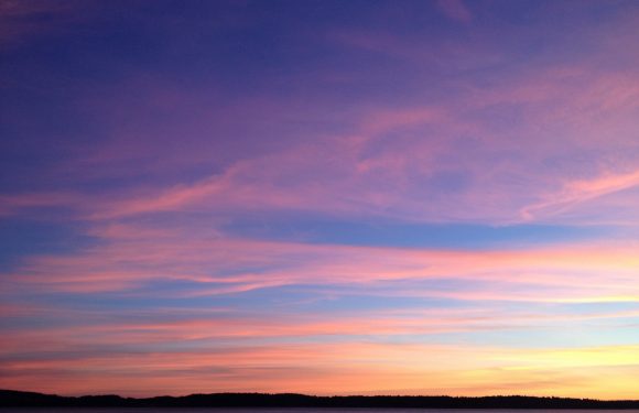A colorful sunset over a calm body of water with a dark silhouette of distant land. The sky features pink, purple, and orange hues with wispy clouds.