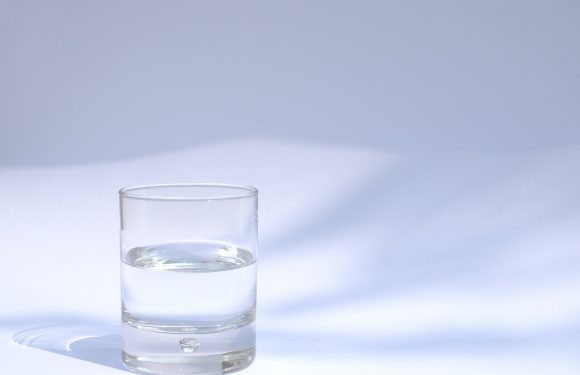 A clear glass of water on a white surface with a subtle shadow, against a light background.