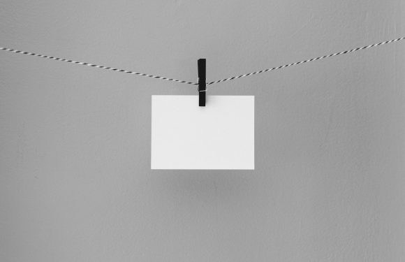 A blank white note card clipped to a twisted black and white string, hanging against a plain light gray wall.