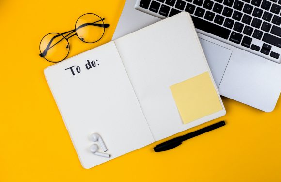 Open notebook with "To do:" written on the left page, a yellow sticky note on the right, a black pen below, white earbuds on the bottom left, a yellow background on the left, and part of a laptop keyboard on the top right.