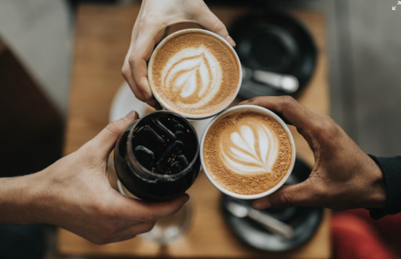 Three people clink glasses: two holding cups of coffee with latte art and one holding a glass of dark soda, over a wooden table.