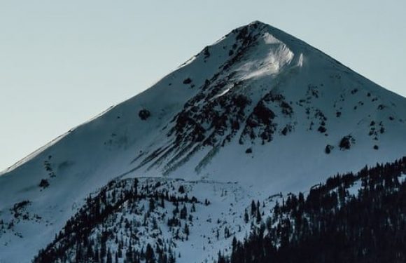 Snow-covered mountain with a clear sky, forest at the base, and patches of snow on the slopes.