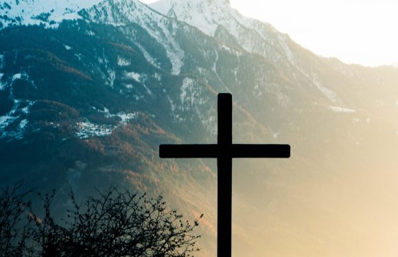 A silhouette of a cross stands in front of misty, snow-capped mountain peaks during sunrise or sunset, with tree branches in the foreground.