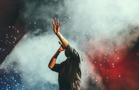 Person on stage with one hand raised, surrounded by colorful smoke or fog in blue, red, and green hues.