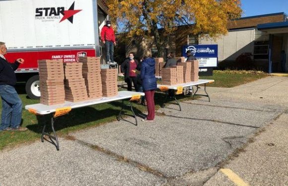 People are unloading pizza boxes onto tables near a large truck with "STAR" written on it. The scene is outdoors with trees and a building in the background.