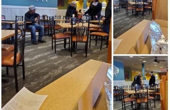 Empty dining area with a blue wall and Pizza Ranch sign, a few staff members serving or preparing tables, and some table settings and foil-wrapped items on the table.