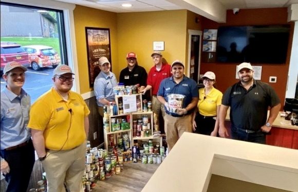 Group of eight employees standing around a donation of canned foods inside a restaurant, with a window showing parked cars outside.