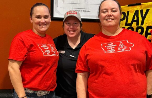 Three women wearing red shirts and a gray cap stand side by side, smiling in front of an orange wall with a black quote board and a promotional sign.