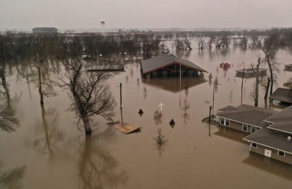 Flooded residential area with submerged houses, trees, and utility poles under gray, overcast sky. Water covers the streets and yards extensively.