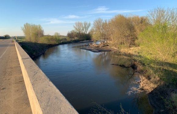 A calm river running alongside a highway with a concrete barrier; trees with early spring foliage line the riverbanks under a blue sky.
