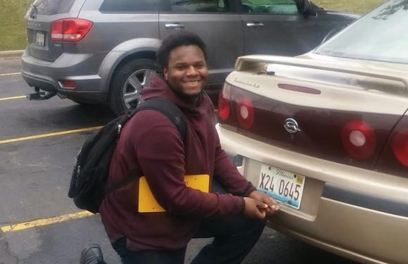 A man kneeling next to a beige car with a black and red spoiler, holding a yellow item, smiling, in a parking lot with other vehicles and a brick building in the background.