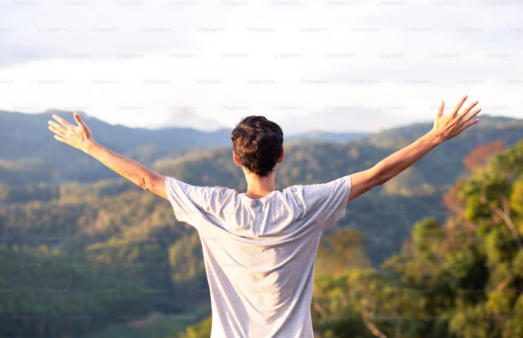 A person standing outdoors with arms outstretched, facing away, overlooking a green mountainous landscape under a cloudy sky.